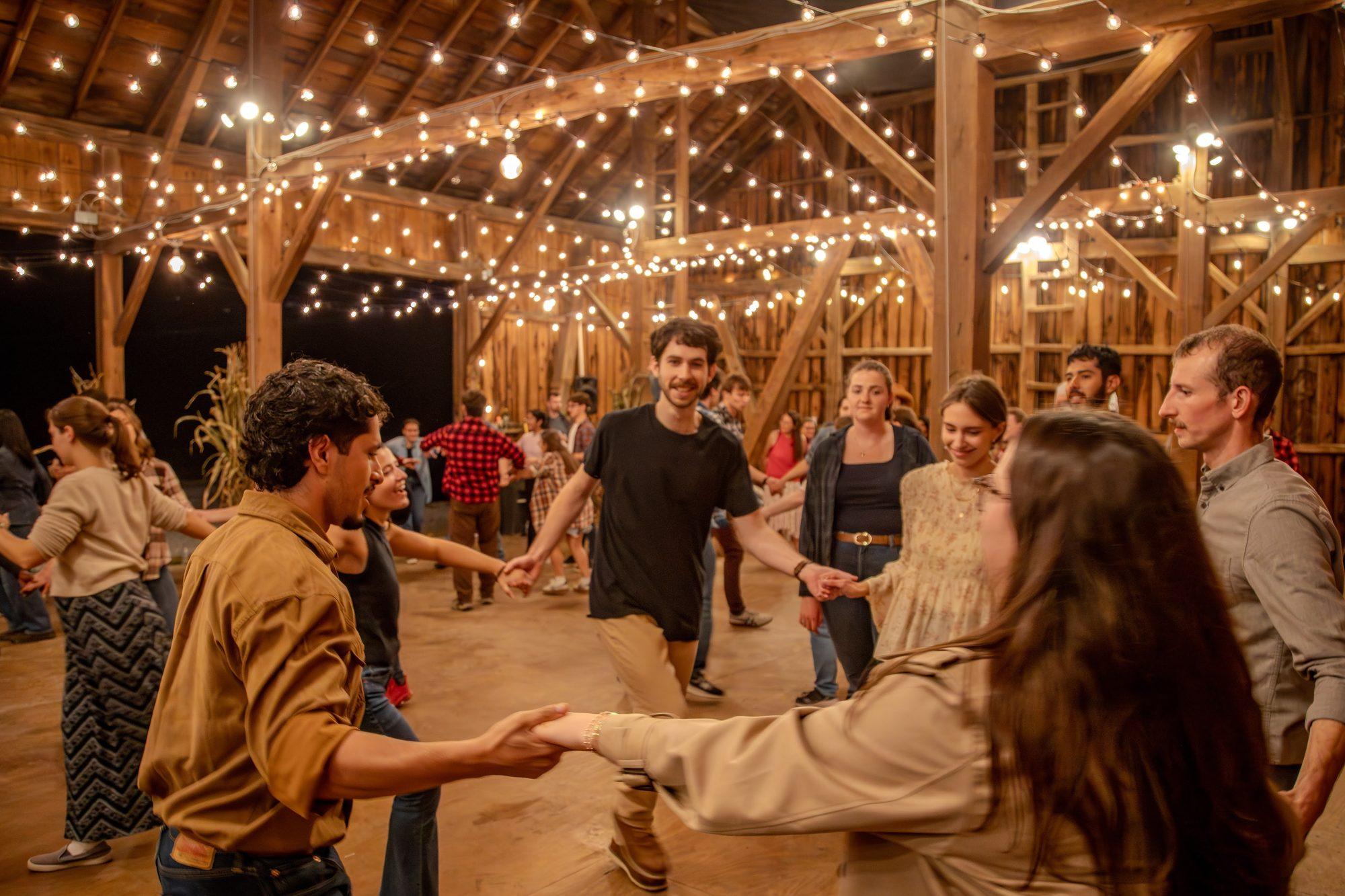 A barn dance circle of people with joined hands under string lights in the Rednal barn